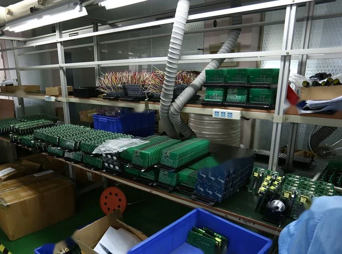 Electronics workshop shelves stacked with green circuit boards, wire harnesses, cardboard boxes, and blue plastic bins