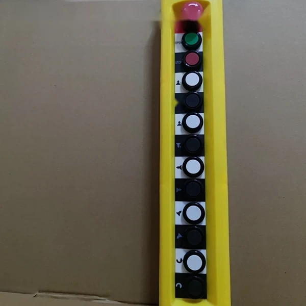 Yellow vertical industrial pendant control station with green, red, white and black push buttons photographed against a cardboard box