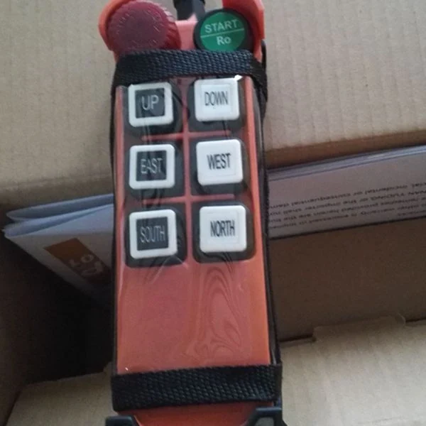 Orange industrial crane remote control with UP, DOWN, EAST, WEST, SOUTH, NORTH buttons, green START button and black strap on a cardboard box