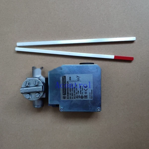 Industrial limit switch with a metal housing and two flat rods, one with a red tip, laid on a brown surface