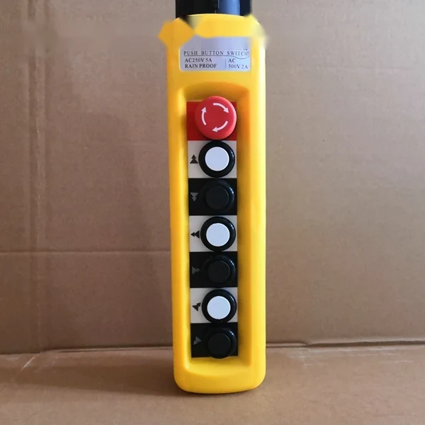 Yellow crane pendant push button switch with red emergency stop and black directional control buttons, on a cardboard box background