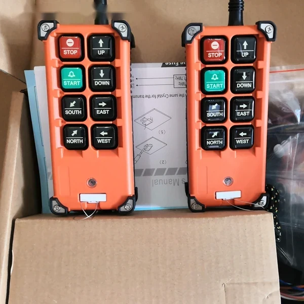 Two orange industrial radio remote controls with STOP, START, UP, DOWN, NORTH, SOUTH, EAST, WEST buttons in a cardboard box