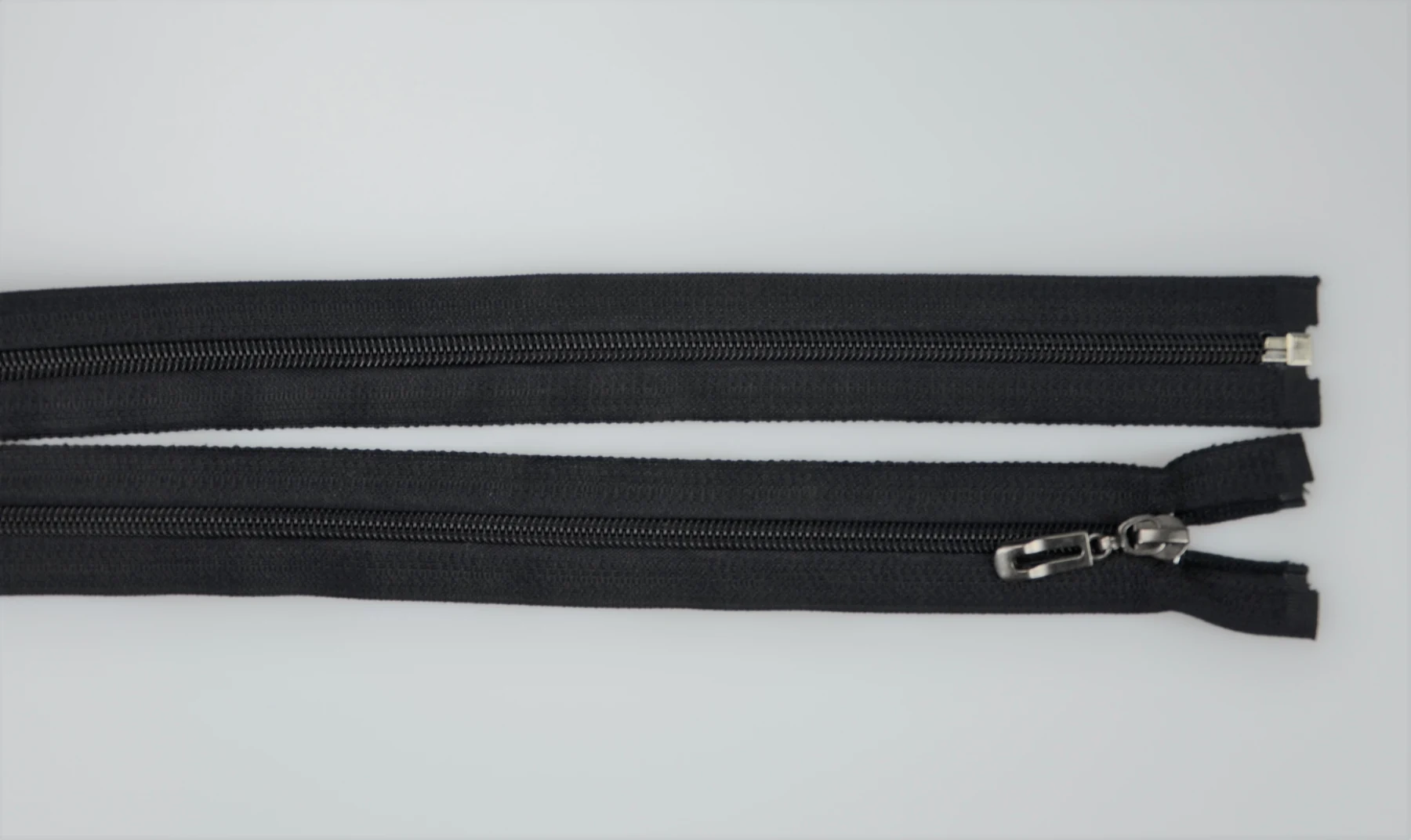 Two black coil zippers on a light gray surface, one with a metal slider and pull, the other detached at one end