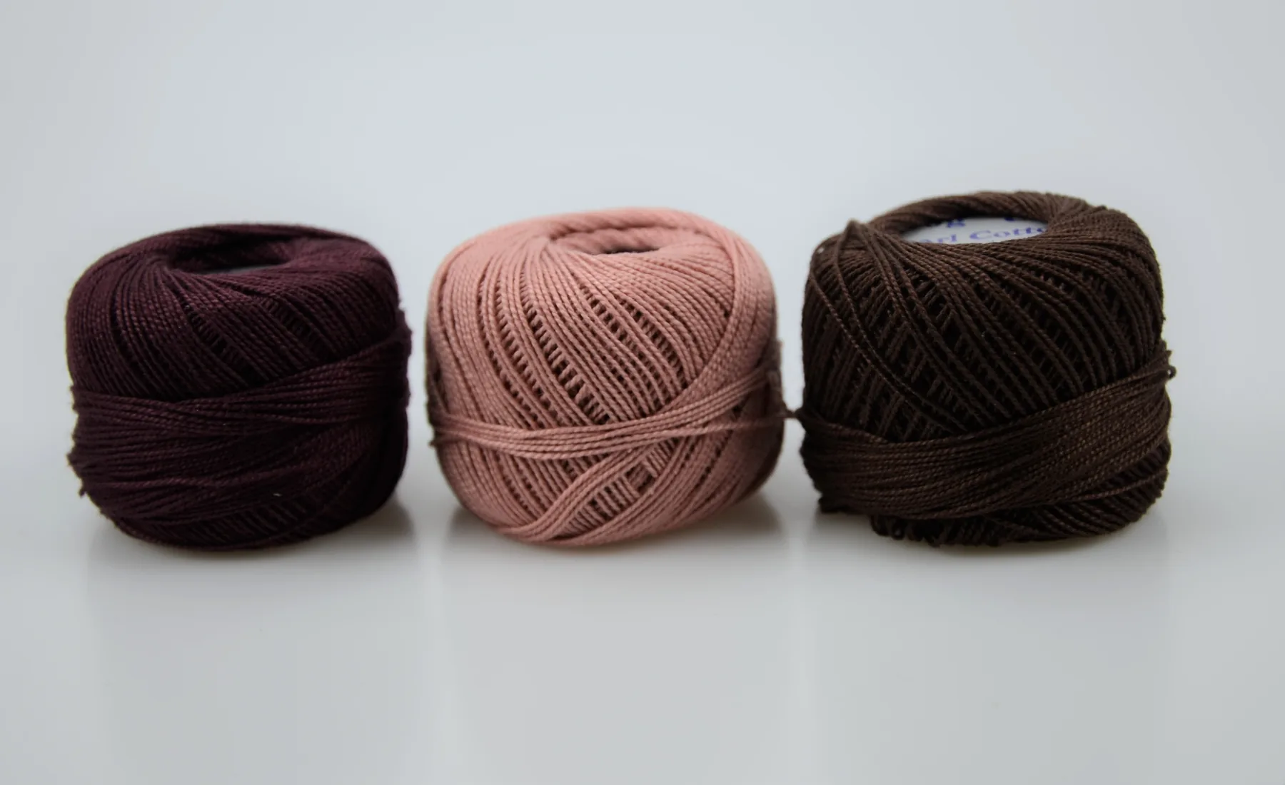 Three balls of yarn in dark burgundy, pink, and dark brown arranged side by side on