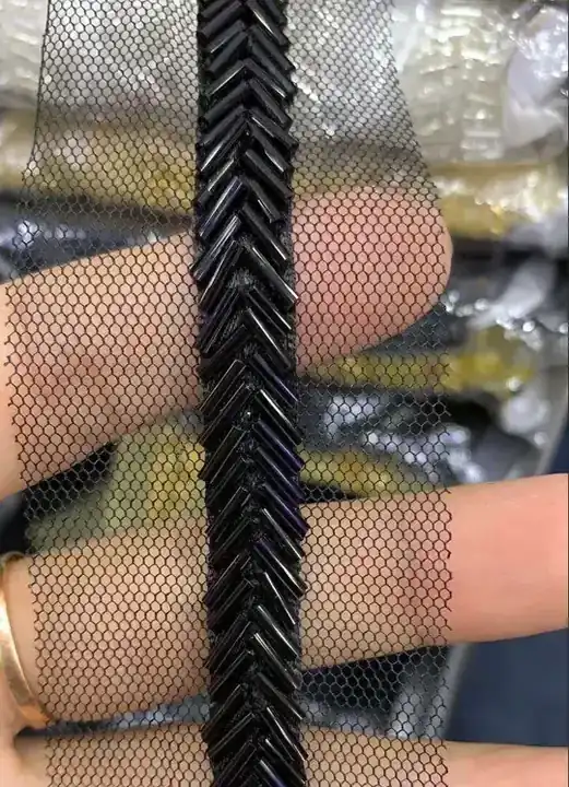 Black bugle bead chevron embroidery on mesh tulle trim, held between fingers, sequin bags blurred