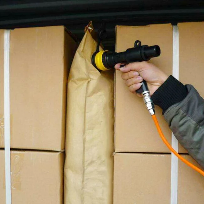 A worker uses a handheld air nozzle to inflate a dunnage bag between cardboard boxes.
