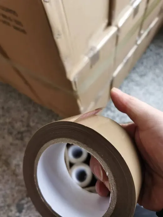 Hand holding a roll of brown kraft packing tape, with stacked cardboard boxes on a