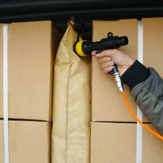 A worker uses a handheld air nozzle to inflate a dunnage bag between cardboard boxes.