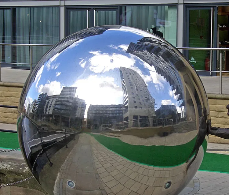 A large, polished silver sphere reflecting a distorted city skyline, buildings, and a blue sky with clouds.