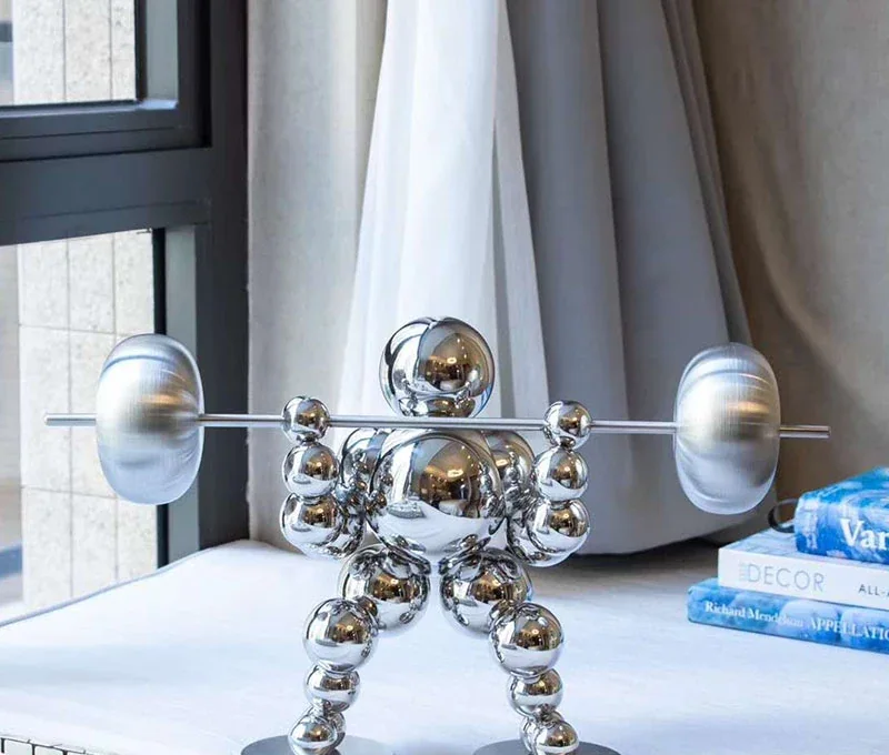 Polished chrome figurine built from stacked spheres lifting a barbell, styled on a white surface beside books