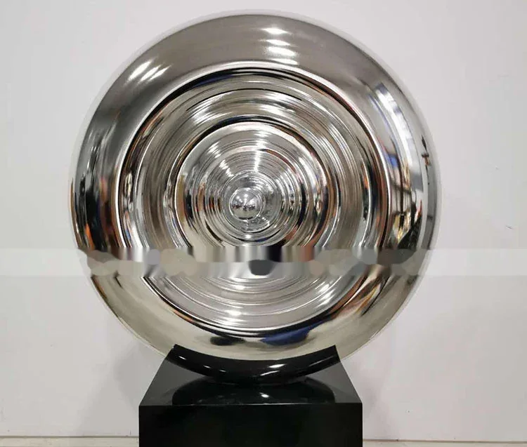 Polished stainless steel disc sculpture with concentric rings mounted on a black square base against a white wall