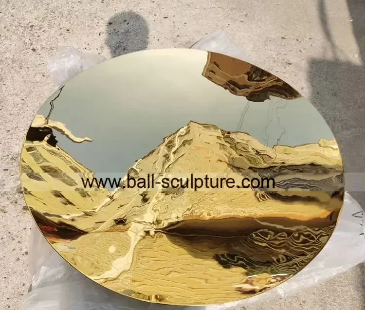 Large polished gold mirror-finish metal disc resting on concrete ground, with a website URL watermarked across it