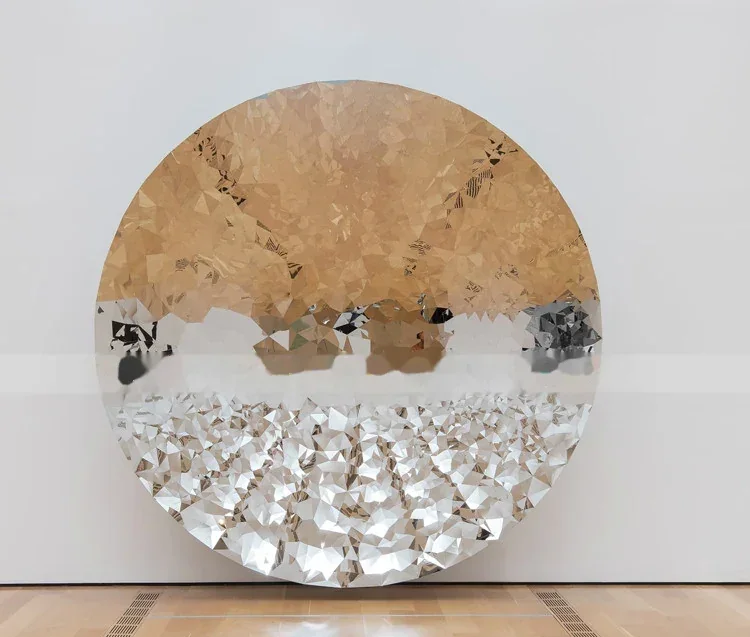 A large circular faceted metal sculpture leaning against a white wall in a gallery.
