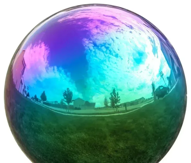Iridescent reflective sphere mirroring sky, clouds, trees and buildings against a plain white background