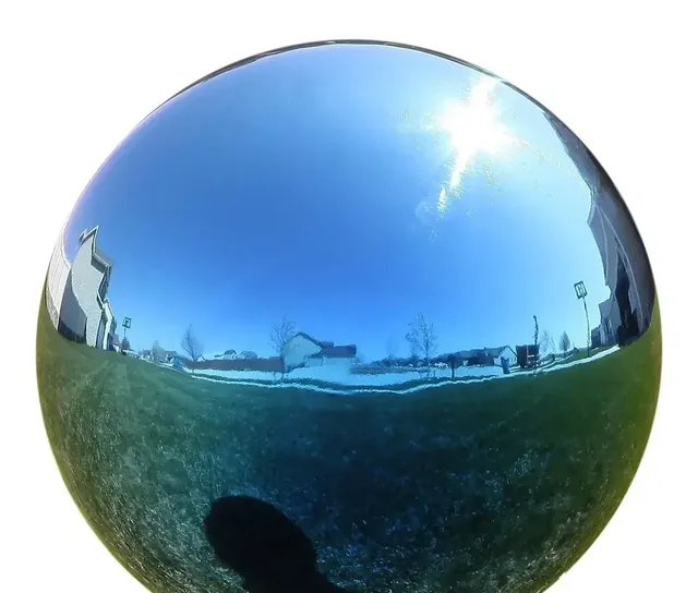 Mirror-polished stainless steel sphere on grass reflecting sky, sun, houses and photographer's shadow
