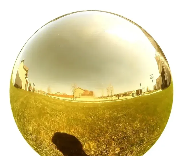 Polished gold mirror-finish sphere photographed outdoors, reflecting grass, buildings, sky and the photographer's shadow