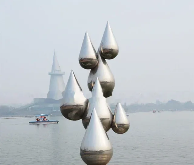 Stainless steel teardrop sculpture by a lake with a boat and distant tower in the background