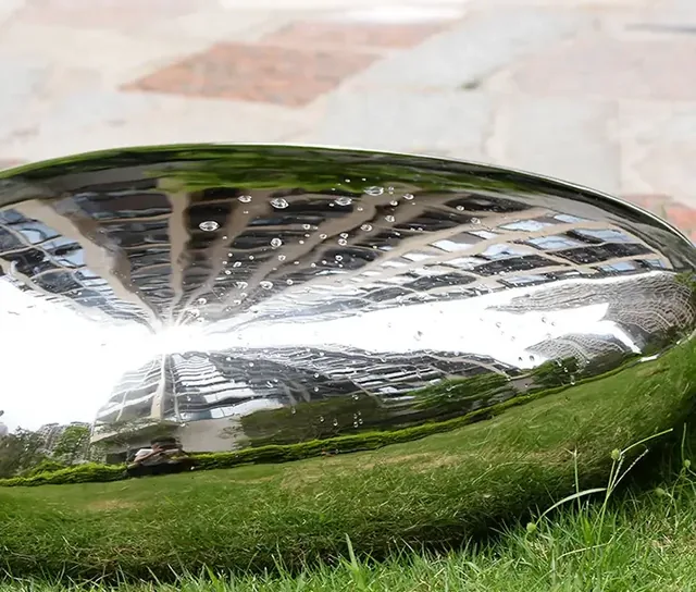 Mirror-polished stainless steel dome on grass, reflecting buildings, trees and sky, with water droplets on its surface