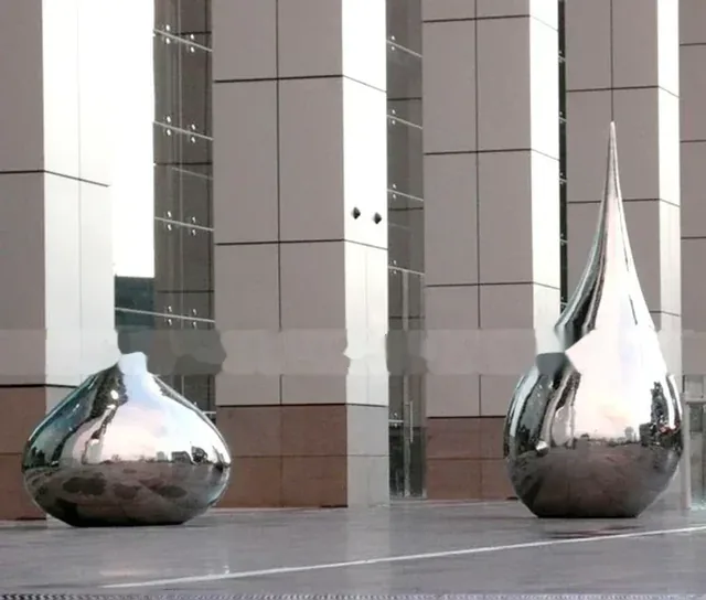 Two large, polished stainless steel sculptures shaped like water droplets stand before a modern building.