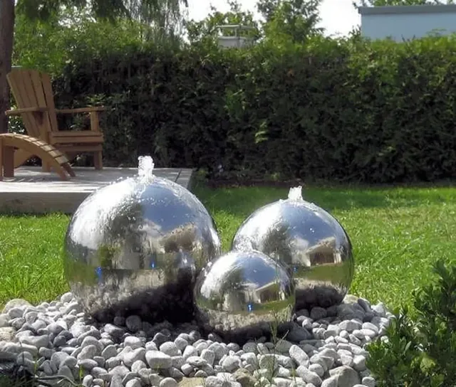Three polished metal spheres with water jets spraying from the top, set on a bed of stones.