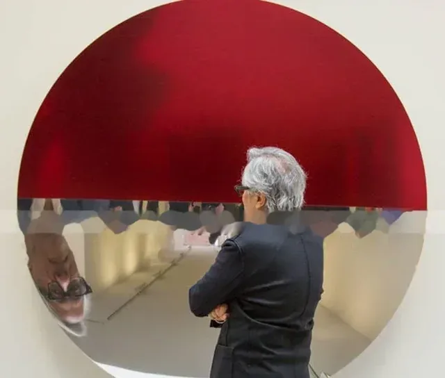 Gray-haired man in dark blazer stands before a large circular artwork with red upper half and mirrored lower half reflecting a gallery interior.