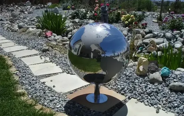 Polished stainless steel globe sculpture on a pedestal in a garden with stone path, gravel, and flowers.