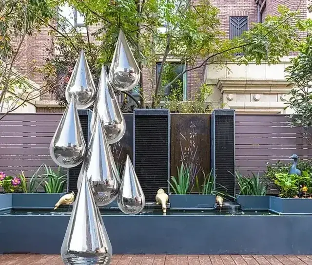 Polished stainless steel teardrop sculptures clustered in a landscaped garden with planters, water feature and brick buildings behind