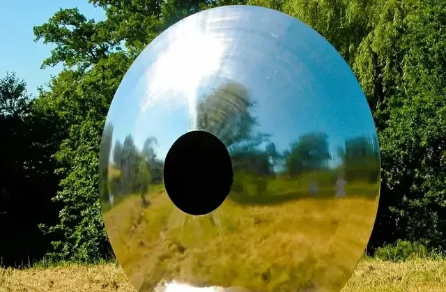 A large, highly polished metal disc with a central hole stands in a grassy field.