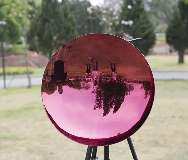 A large, pink, concave mirror on a tripod outdoors, reflecting an inverted view of people and trees.