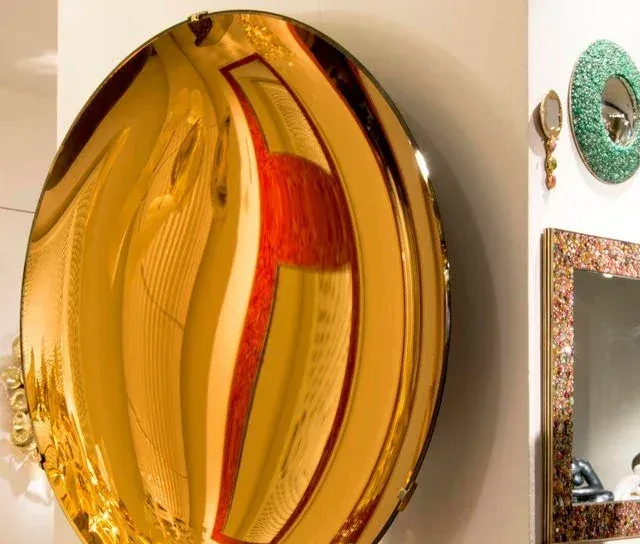 A large, round gold convex mirror mounted on a wall, reflecting a red chair and room interior.