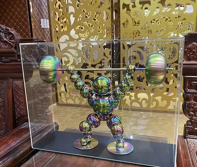 Iridescent metal sculpture of a weightlifter holding a barbell, displayed in a clear acrylic case.