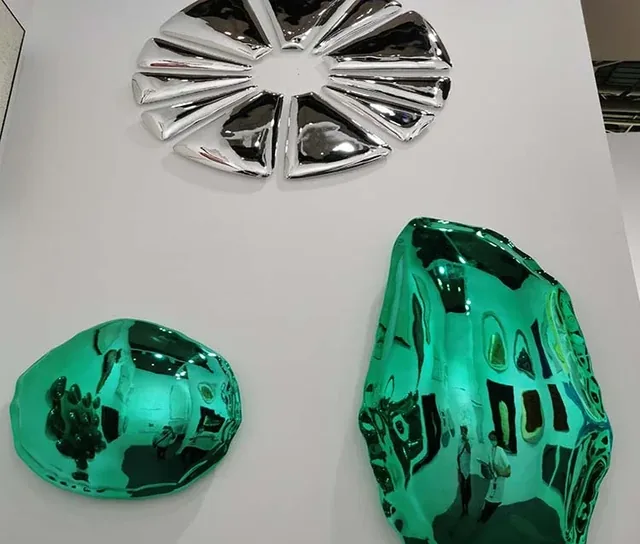 Three metallic sculptures, one silver and two green, with reflective surfaces arranged on a white display surface.