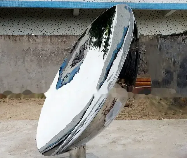 Large polished stainless steel disc on a stand photographed outdoors against a weathered concrete wall