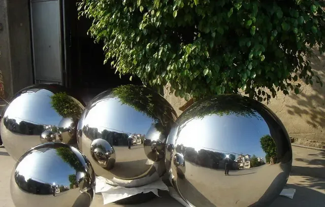 Polished stainless steel spheres of various sizes displayed outdoors under a leafy tree
