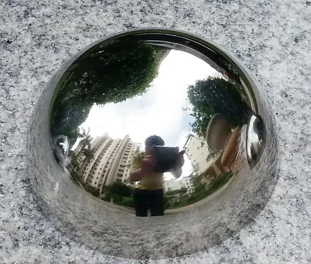 Polished stainless steel hemisphere dome on a granite surface, its mirror finish reflecting buildings, trees, and sky
