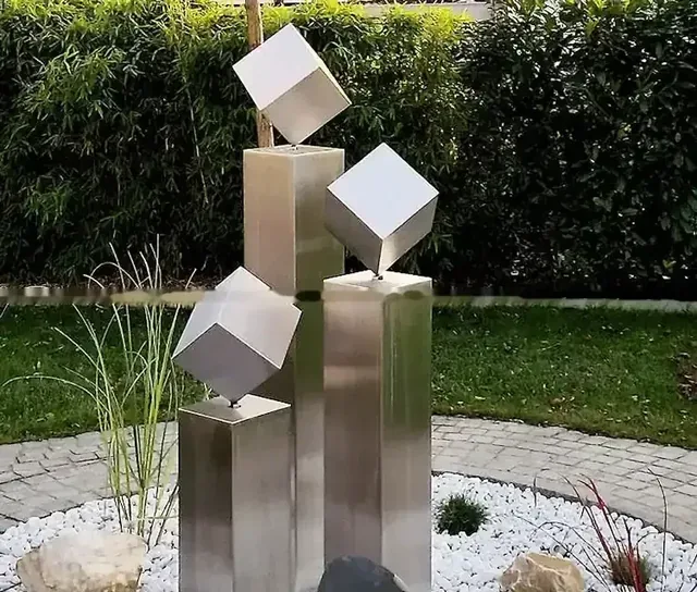 Modern stainless steel geometric sculpture featuring balanced cubes on pillars in a garden