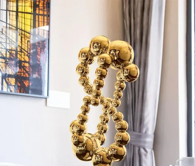 Polished golden sculpture made of clustered reflective spheres, shown indoors beside a curtain and framed wall art.