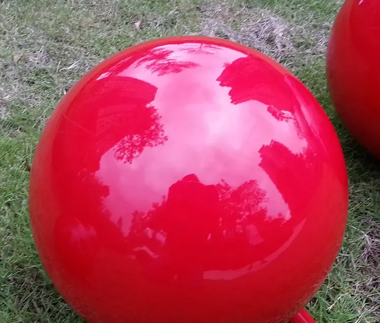 Large glossy red sphere resting on grass, tree and sky reflections on its surface, second sphere partly visible