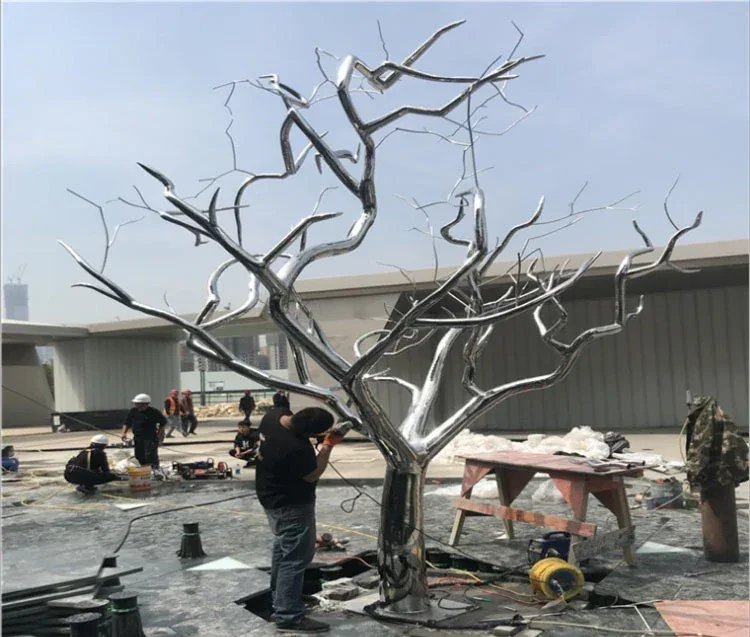 Workers assembling a large polished stainless steel tree sculpture at an outdoor construction site.