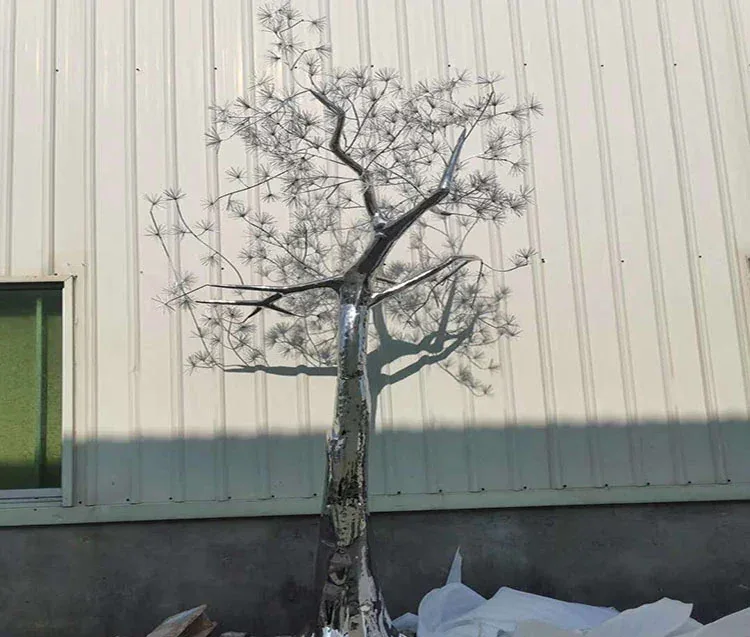 Polished stainless steel tree sculpture standing against a corrugated metal warehouse wall