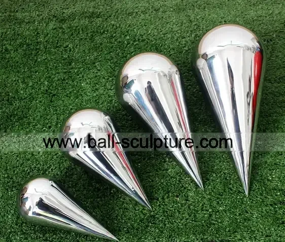 Four polished stainless steel teardrop spike sculptures in ascending sizes lying on green artificial grass