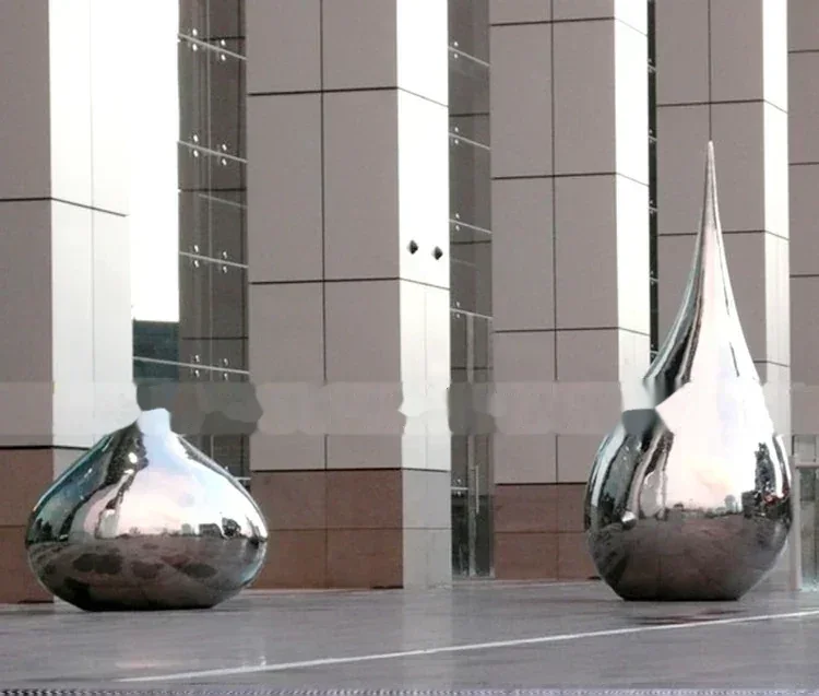 Two large, polished stainless steel sculptures shaped like water droplets stand before a modern building.