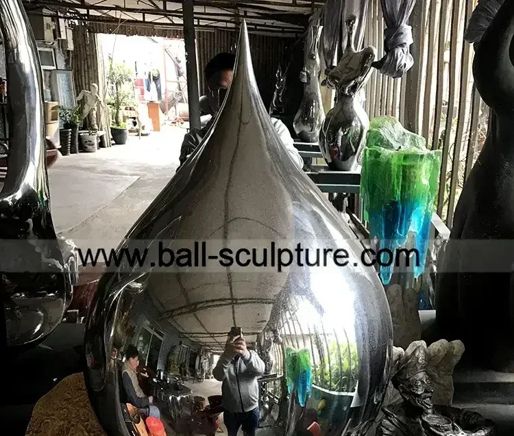 Polished stainless steel teardrop sculpture in a workshop surrounded by other metal artworks