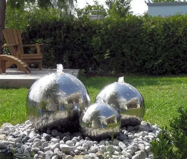 Three polished metal spheres with water jets spraying from the top, set on a bed of stones.