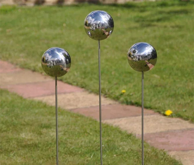 Three polished stainless steel spheres on thin metal stakes displayed in a lawn beside a stone pathway