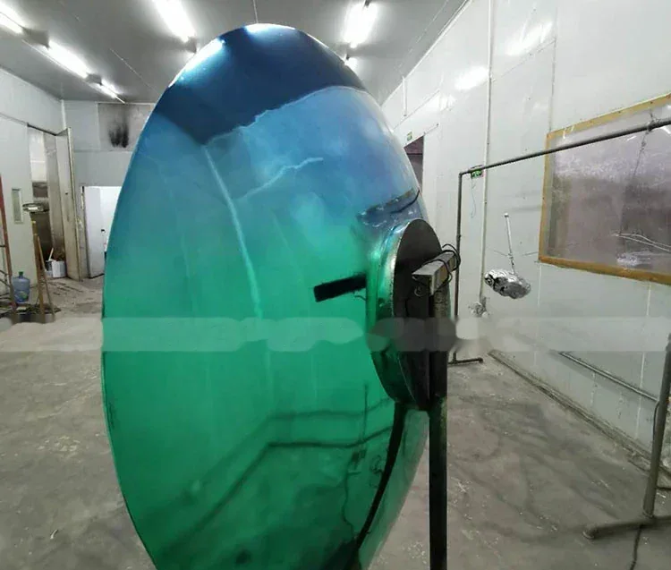 Large oval curved glass panel with blue and green gradient standing on a mount in a workshop.