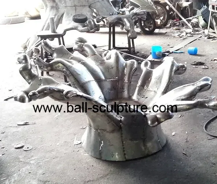 Unfinished welded stainless steel splash sculpture on a cluttered workshop floor, URL burned across image