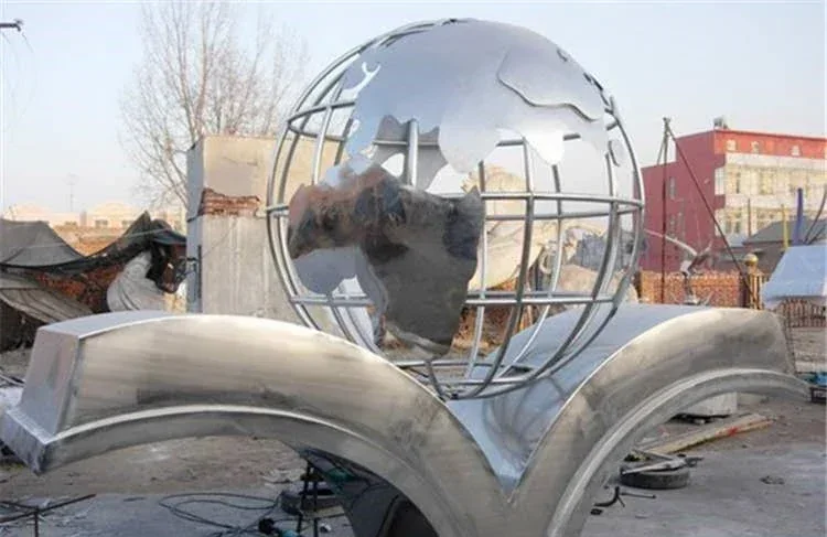 Polished stainless steel globe sculpture on an open book base in an outdoor fabrication yard