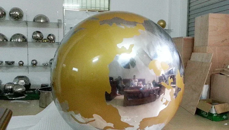 Large mirror-polished steel sphere sculpture with gold-toned continents, photographed in a workshop with shelved steel spheres behind