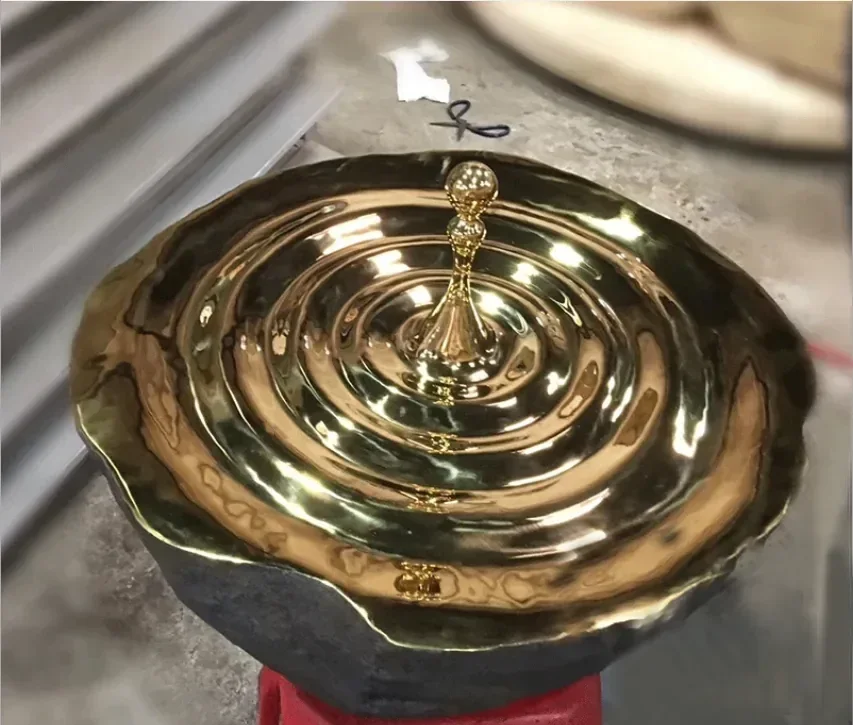 Polished brass sculpture shaped like a water droplet with concentric ripple rings, photographed in a workshop